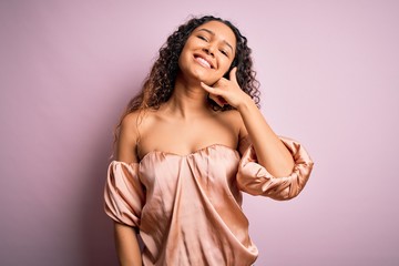 Young beautiful woman with curly hair wearing casual t-shirt standing over pink background smiling doing phone gesture with hand and fingers like talking on the telephone. Communicating concepts.