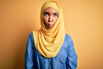 Young beautiful woman with curly hair wearing arab traditional hijab over yellow background making fish face with lips, crazy and comical gesture. Funny expression.