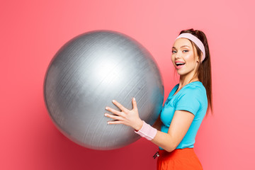 Fototapeta premium cheerful sportswoman holding silver fitness ball while smiling at camera on pink background