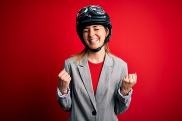 Young beautiful blonde motorcyclist woman wearing motorcycle helmet over red background excited for success with arms raised and eyes closed celebrating victory smiling. Winner concept.