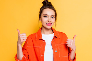 attractive girl showing thumbs up while looking at camera isolated on yellow