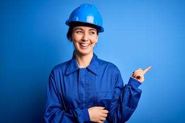 Young beautiful worker woman with blue eyes wearing security helmet and uniform with a big smile on face, pointing with hand and finger to the side looking at the camera.