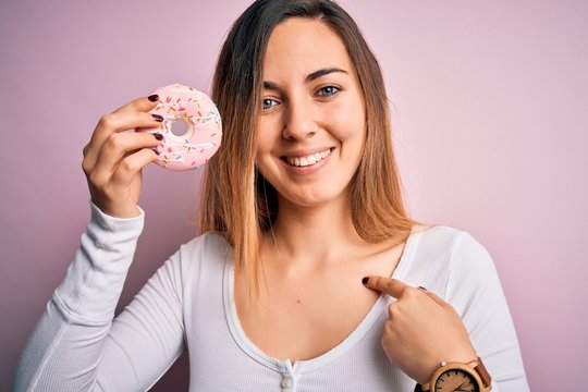 Young beautiful blonde woman with blue eyes holding pink doughnut over isolated background with surprise face pointing finger to himself