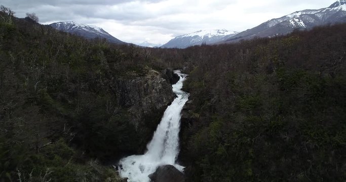 Aerial flying towards and above waterfall between dense woods at winter, discovering plain valley of winter trees structure. Snowy Andes mountain chains at background. Vullignanco fall, Nequen