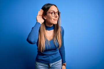 Young beautiful blonde woman with blue eyes wearing glasses standing over blue background smiling with hand over ear listening an hearing to rumor or gossip. Deafness concept.