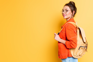 attractive, smiling student with backpack looking at camera on yellow background