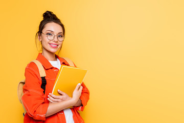 cheerful student in glasses looking at camera while holding notebook isolated on yellow - obrazy, fototapety, plakaty