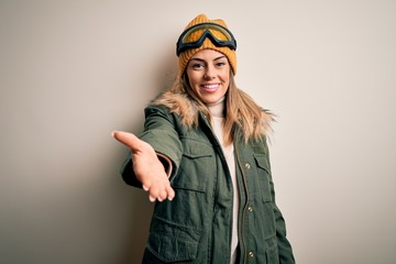 Young brunette skier woman wearing snow clothes and ski goggles over white background smiling cheerful offering palm hand giving assistance and acceptance.