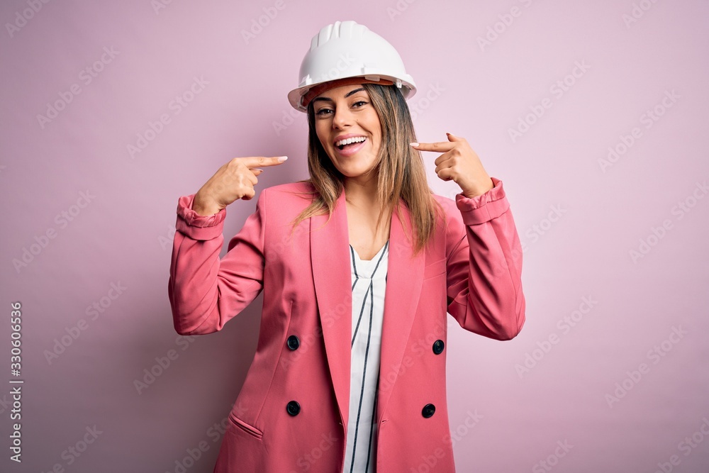 Poster young beautiful brunette architect woman wearing safety helmet over pink background smiling cheerful