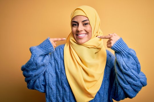 Young Beautiful Brunette Muslim Woman Wearing Arab Hijab Over Isolated Yellow Background Smiling Cheerful Showing And Pointing With Fingers Teeth And Mouth. Dental Health Concept.