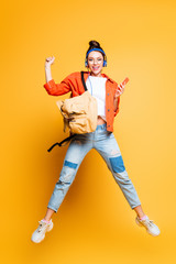 cheerful student in headset and glasses jumping while holding smartphone on yellow background