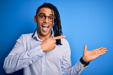 Young handsome african american man with dreadlocks wearing casual shirt and glasses amazed and smiling to the camera while presenting with hand and pointing with finger.