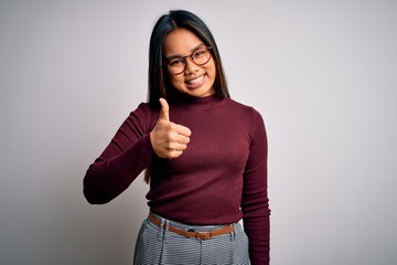 Beautiful asian business woman wearing casual sweater and glasses over white background doing happy thumbs up gesture with hand. Approving expression looking at the camera showing success.