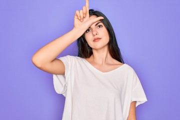 Obraz na płótnie Canvas Young beautiful brunette woman wearing casual white t-shirt over purple background making fun of people with fingers on forehead doing loser gesture mocking and insulting.