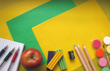 Top view of school stationery: paints, colored pencils, pens, plasticine, notepad and apple on a background of colored paper. Back to school flat lay with copy space