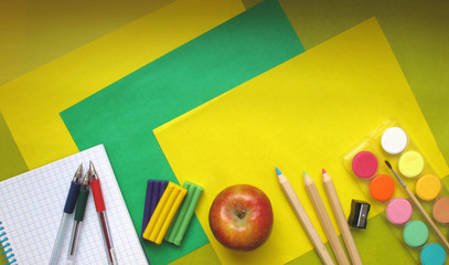 Top view of school stationery: paints, colored pencils, pens, notepad and apple on a background of colored paper. Back to school flat lay with copy space