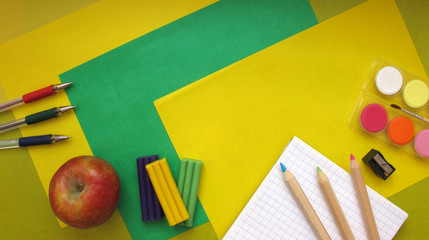 Top view of school stationery: paints, colored pencils, pen, plasticine, notepad and apple on a background of colored paper. Back to school flat lay with copy space