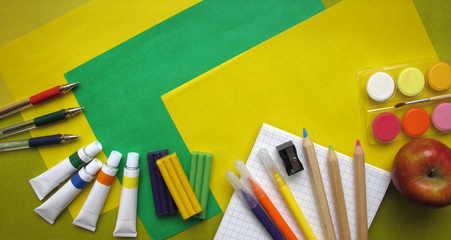 Top view of school stationery: paints, colored pencils, pen, plasticine, notepad and apple on a background of colored paper. Back to school flat lay with copy space