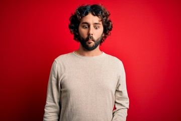 Fototapeta na wymiar Young handsome man with beard wearing casual sweater standing over red background making fish face with lips, crazy and comical gesture. Funny expression.