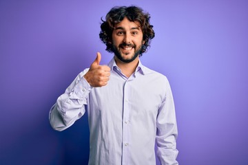 Young handsome business man with beard wearing shirt standing over purple background doing happy thumbs up gesture with hand. Approving expression looking at the camera showing success.