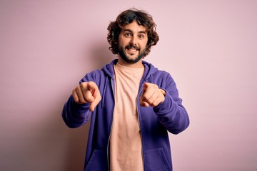 Young handsome sporty man with beard wearing casual sweatshirt over pink background pointing to you and the camera with fingers, smiling positive and cheerful