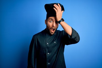 Young handsome chef man with beard wearing cooker uniform and hat over blue background surprised...