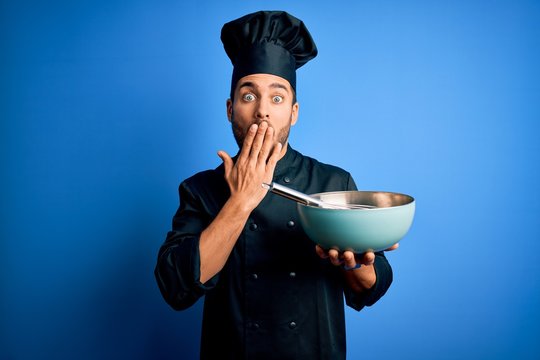 Young cooker man with beard wearing uniform using whisk and bowl over blue background cover mouth with hand shocked with shame for mistake, expression of fear, scared in silence, secret concept