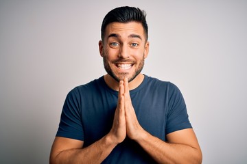 Young handsome man wearing casual t-shirt standing over isolated white background praying with hands together asking for forgiveness smiling confident.