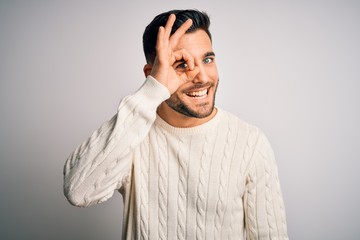Young handsome man wearing casual sweater standing over isolated white background doing ok gesture with hand smiling, eye looking through fingers with happy face.