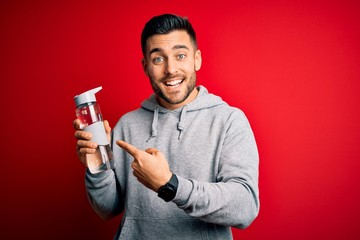 Young handsome sportsman drinking bottle of water to refeshment over red background very happy pointing with hand and finger