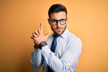 Young handsome businessman wearing tie and glasses standing over yellow background Holding symbolic gun with hand gesture, playing killing shooting weapons, angry face