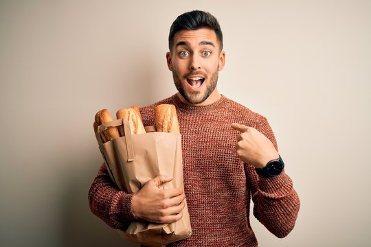 Young handsome man holding groceries paper bag of fresh baguette bread over isolated background with surprise face pointing finger to himself