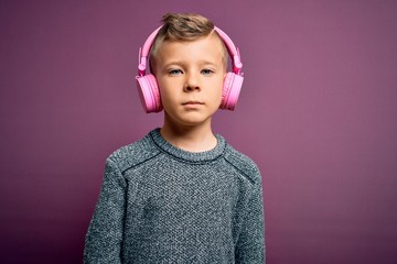 Young little caucasian kid wearing headphones listening to music over purple background with serious expression on face. Simple and natural looking at the camera.