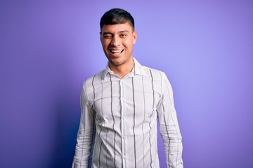 Young handsome hispanic man wearing elegant business shirt standing over purple background winking looking at the camera with sexy expression, cheerful and happy face.