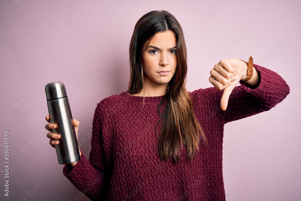 Canvas Prints Young beautiful girl holding thermo with water standing over isolated pink background with angry face, negative sign showing dislike with thumbs down, rejection concept