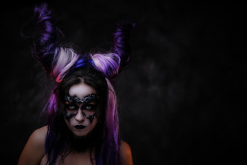 Portrait of a demonic and wild looking girl wearing scary fantasy make up and violet horns posing in a dark studio on a grey background