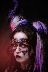 Portrait of a demonic girl wearing scary fantasy make up and violet horns posing in a dark studio with red light on a grey background and looking spooky. Close up view.