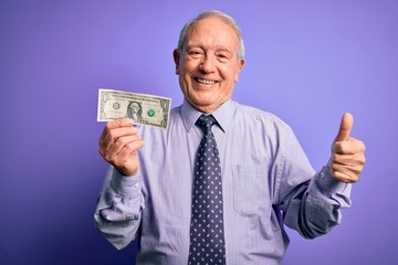 Senior grey haired man holding one dollar bank note over purple background happy with big smile doing ok sign, thumb up with fingers, excellent sign