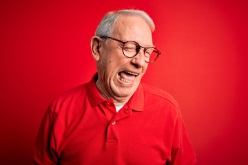 Grey haired senior man wearing glasses and casual t-shirt over red background winking looking at...