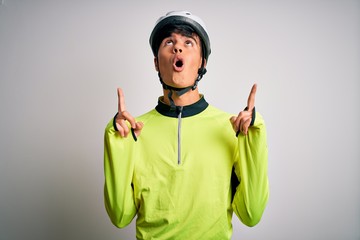 Young handsome cyclist man wearing security bike helmet over isolated white background amazed and surprised looking up and pointing with fingers and raised arms.
