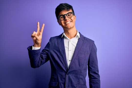 Young handsome business man wearing jacket and glasses over isolated purple background smiling looking to the camera showing fingers doing victory sign. Number two.