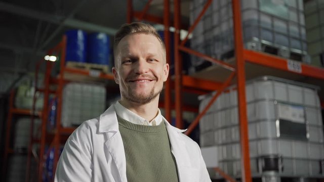 Panning from below portrait of middle aged engineer in white coat looking at camera and smiling standing in industrial storage