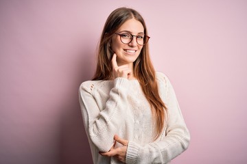 Obraz premium Young beautiful redhead woman wearing casual sweater and glasses over pink background looking confident at the camera smiling with crossed arms and hand raised on chin. Thinking positive.