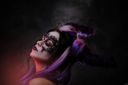 Portrait of a devil girl wearing scary fantasy make up and violet horns posing in a dark studio on a grey background with eyes closed