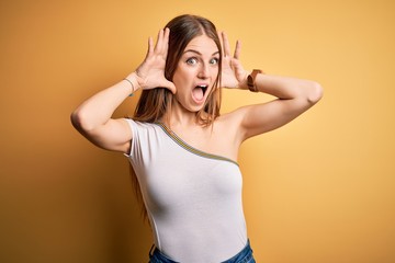 Young beautiful redhead woman wearing casual t-shirt over isolated yellow background Smiling cheerful playing peek a boo with hands showing face. Surprised and exited