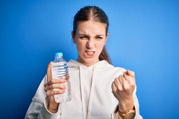 Young beautiful redhead woman doing sport drinking bottle of water over blue background annoyed and frustrated shouting with anger, crazy and yelling with raised hand, anger concept