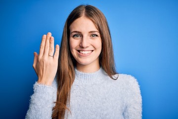 Young beautiful redhead woman wearing wedding ring on finger over blue background with a happy face...