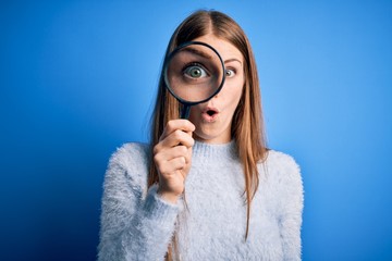 Young beautiful redhead detective woman using magnifying glass over isolated blue background scared...
