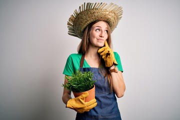 Young beautiful redhead farmer woman wearing apron and hat holding plant pot serious face thinking about question, very confused idea