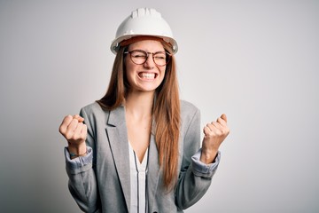 Young beautiful redhead architect woman wearing security helmet over white background very happy and excited doing winner gesture with arms raised, smiling and screaming for success. Celebration
